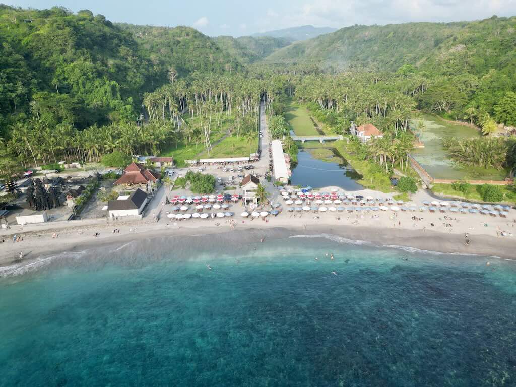 Pemandangan drone Crystal Bay Nusa Penida dengan pantai, pura kecil di tepi laut, dan perbukitan hijau di belakang teluk