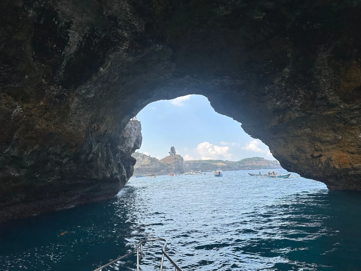 Broken Beach arch seen from inside the bay from a dive boat, with snorkeling boats over Manta Bay and the Octopus Queen sculpture on the headland, Nusa Penida