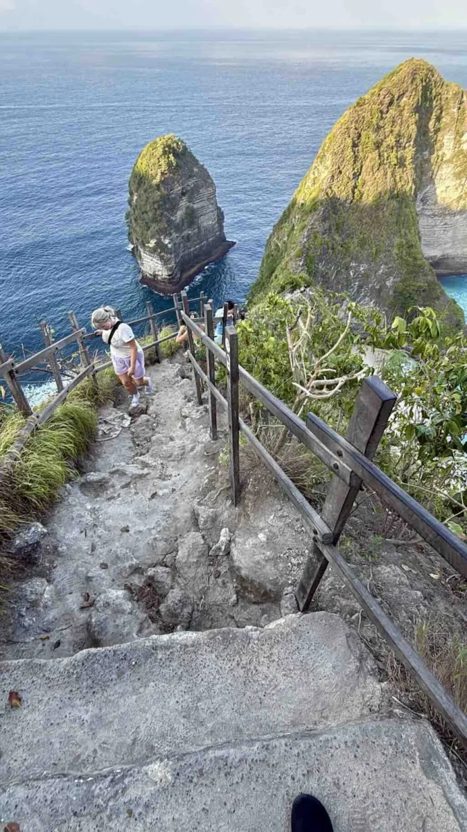 Pandangan dari atas ke anak tangga jalur turun Kelingking Beach yang sudah aus, dengan pegangan kayu, seorang pendaki yang sedang naik, dan batu karang serta laut di kejauhan