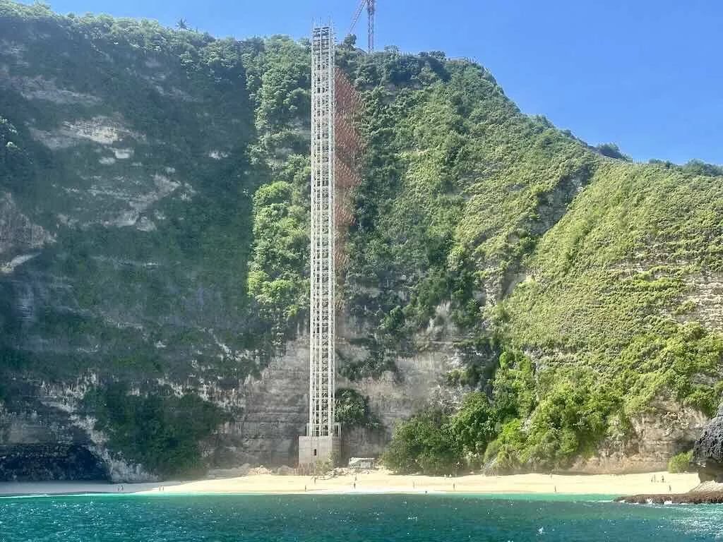 Rangka lift yang belum selesai menjulang di tebing Kelingking Beach, dilihat dari perahu, dengan pantai berpasir putih di bawahnya