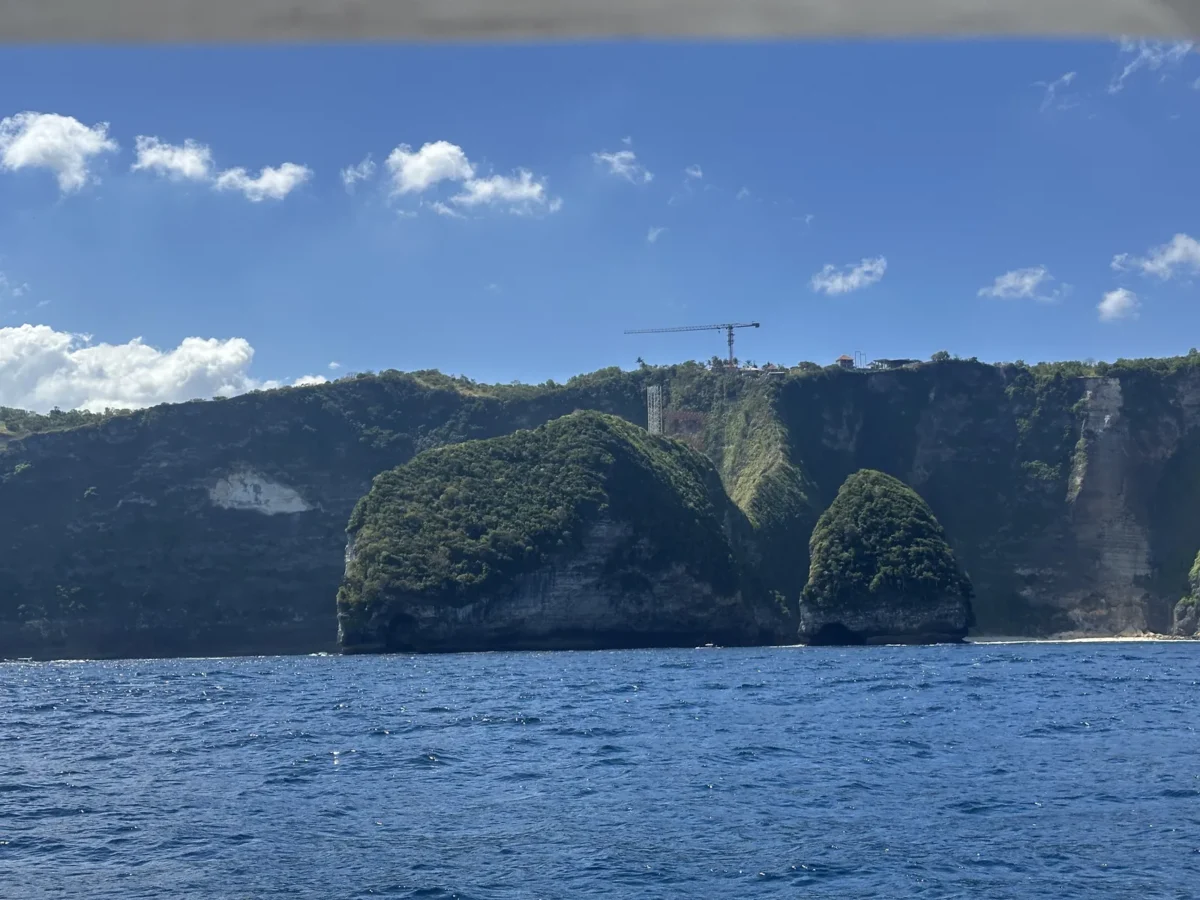 Tanjung Kelingking dilihat dari kejauhan di laut, dengan crane proyek yang masih berdiri di atas tebing di atas rangka lift